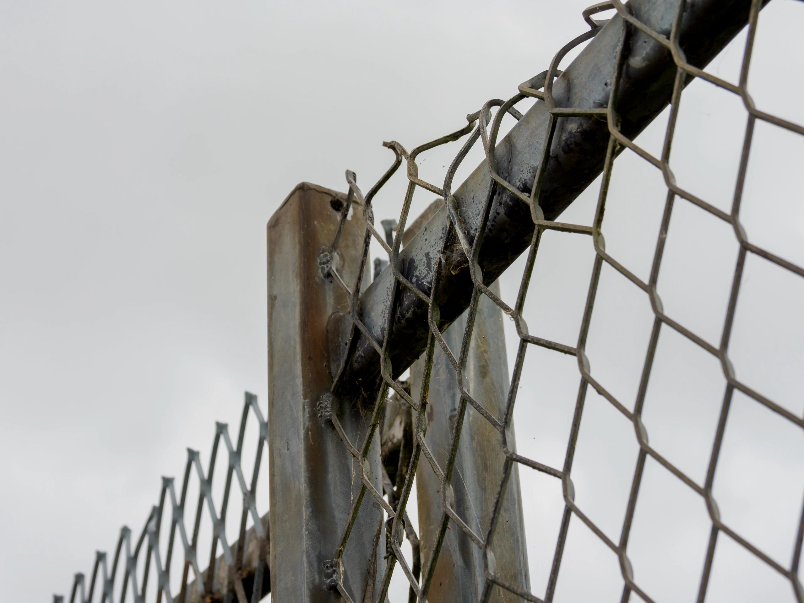 Close-up of a weathered chain-link fence with a broken section under an overcast sky.