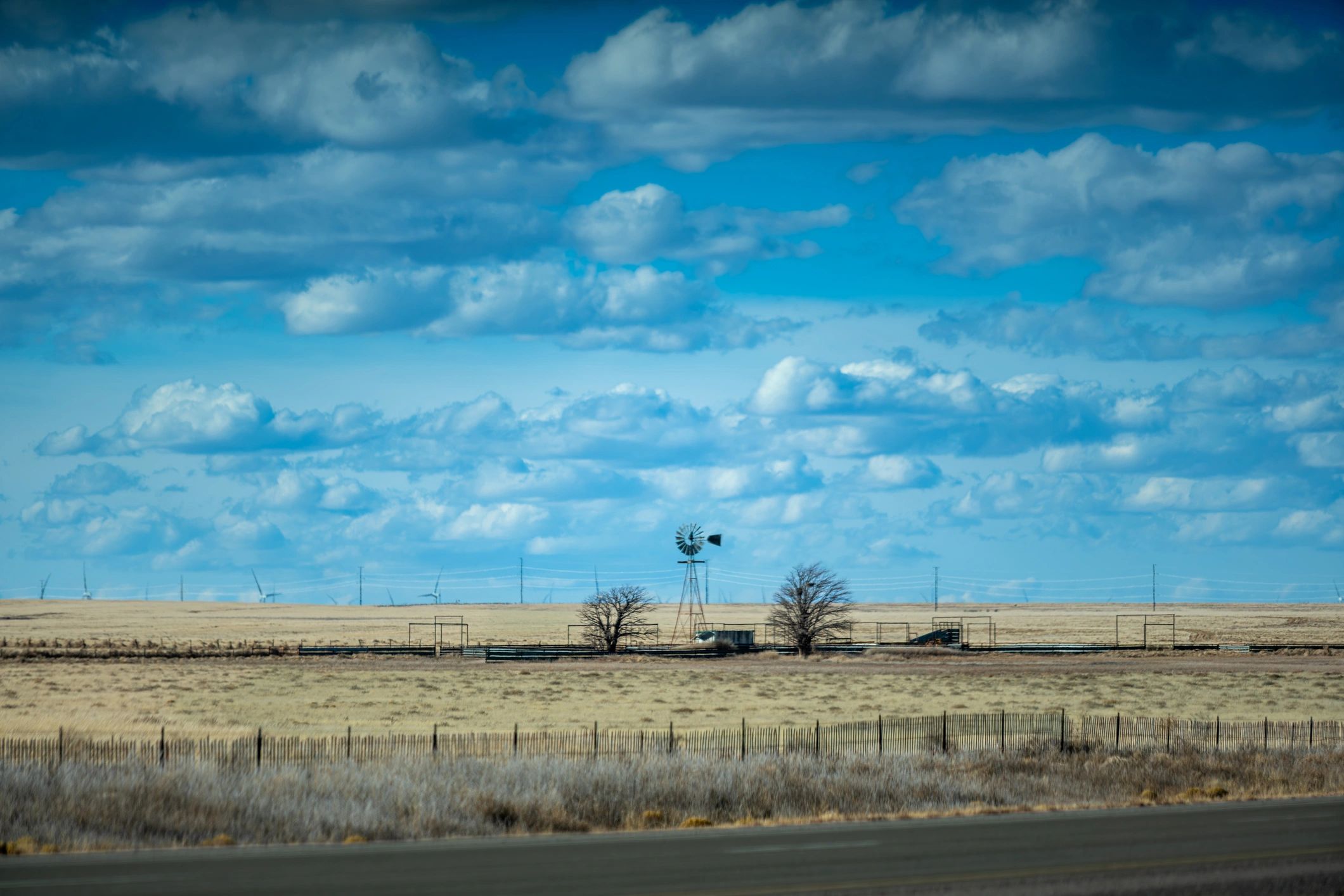 Rural ranch property with windmill and livestock fencing across high plains grassland. Sparse vegetation and arid landscape under clear blue sky.